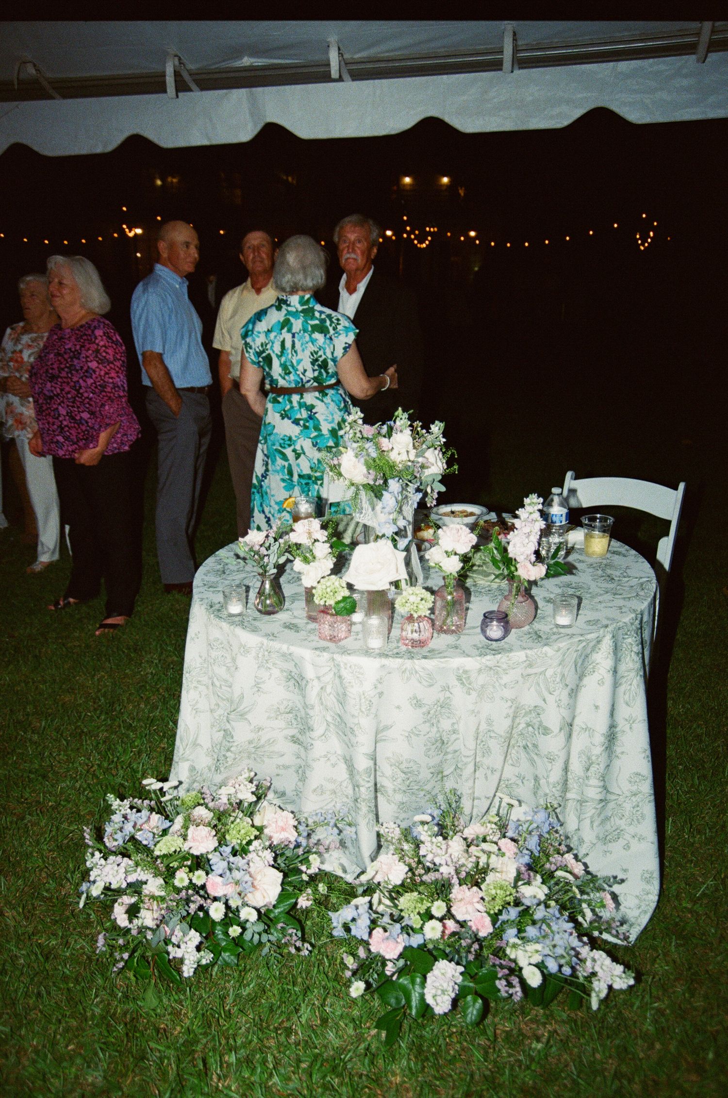 Wedding reception table decorated with white linens and floral arrangements illuminated by string lights at night.