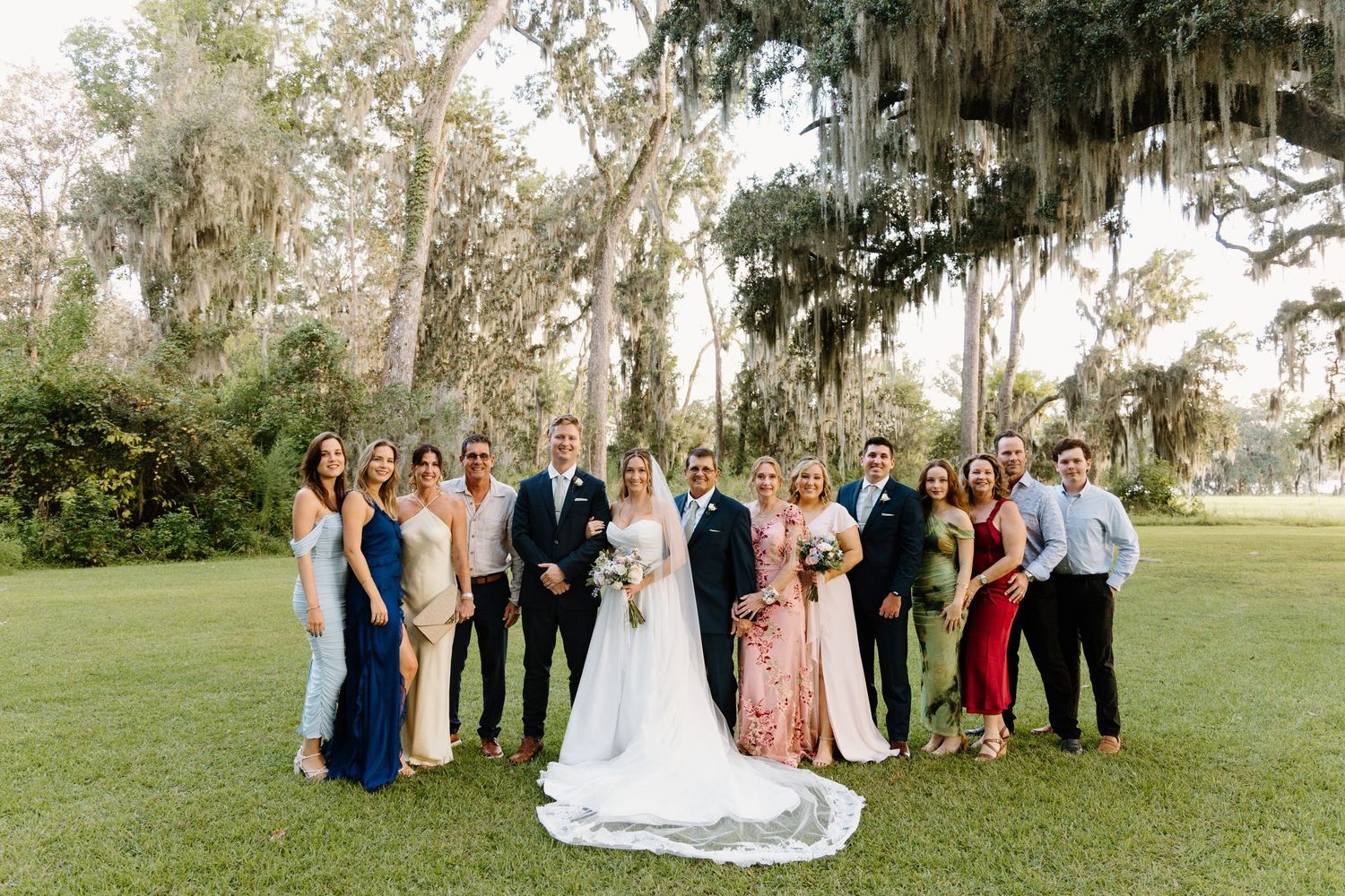 Wedding party poses on a lush green lawn under Spanish moss-draped trees at an elegant outdoor venue.