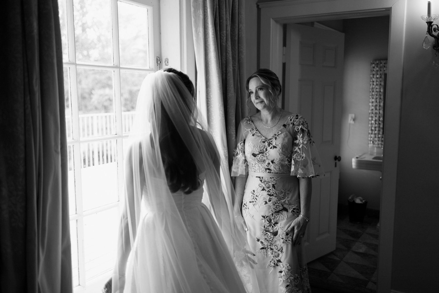 Black and white photo of a wedding dress and veil hanging by a window in soft natural light.