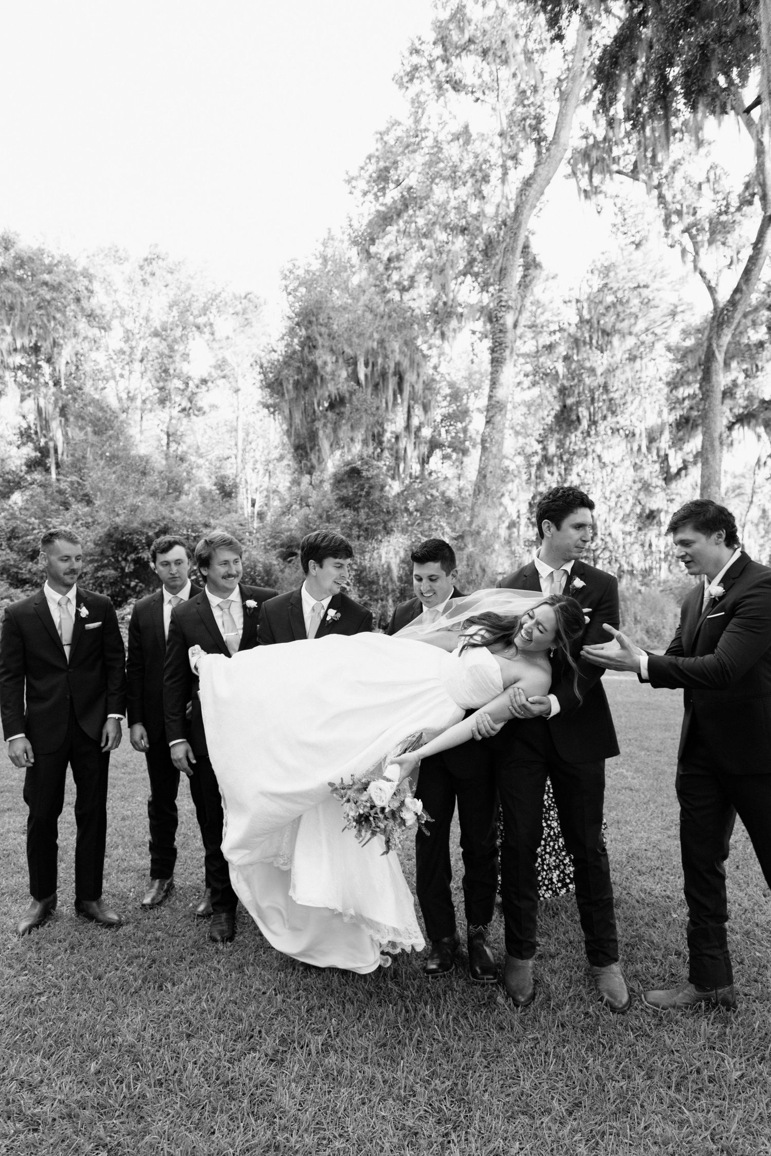 Wedding party lined up outdoors in black and white photo with flowing dress and trees in background.