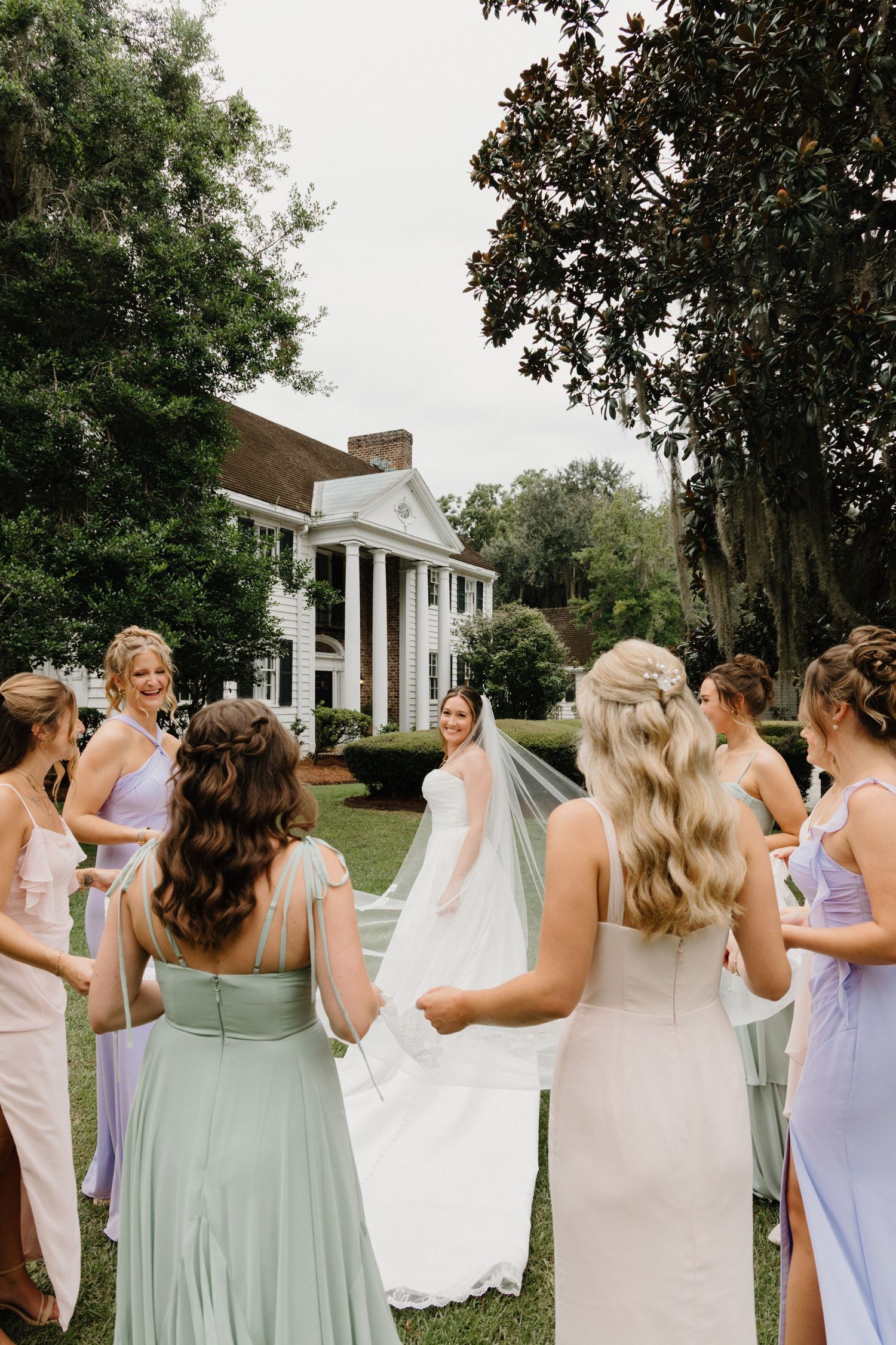 Bridesmaids in pastel purple and mint dresses gather around a bride in white against a colonial-style building backdrop.