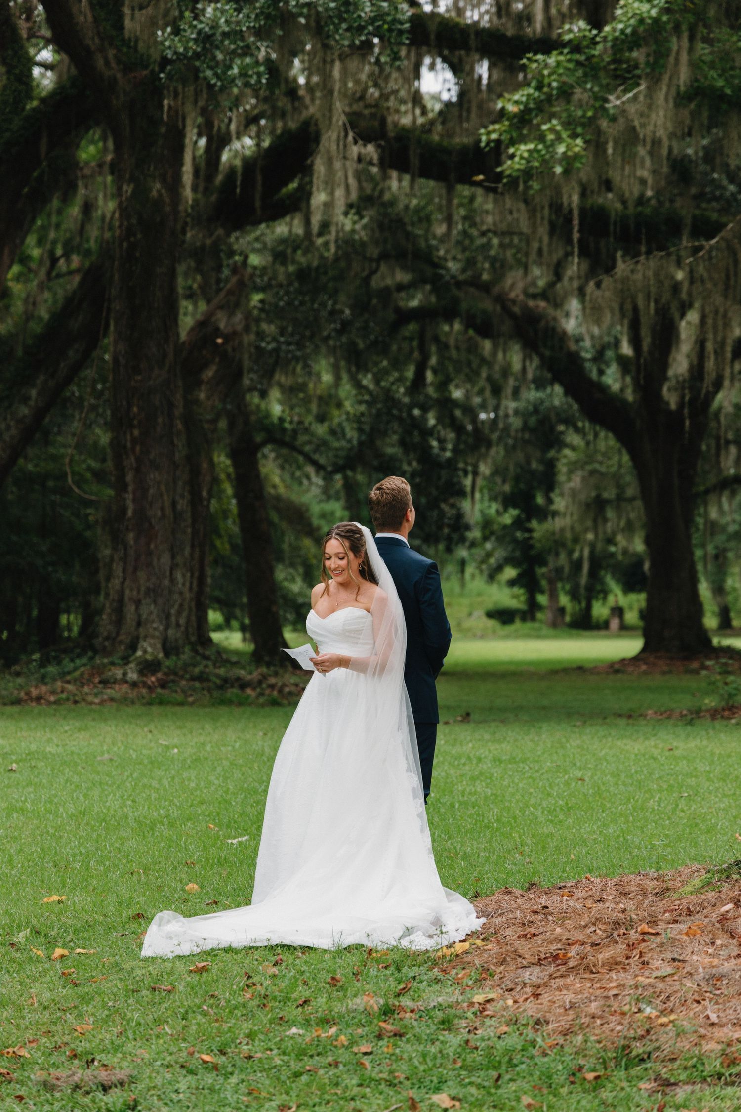 A couple in wedding attire sits together on a grassy lawn surrounded by Spanish moss draped oak trees.