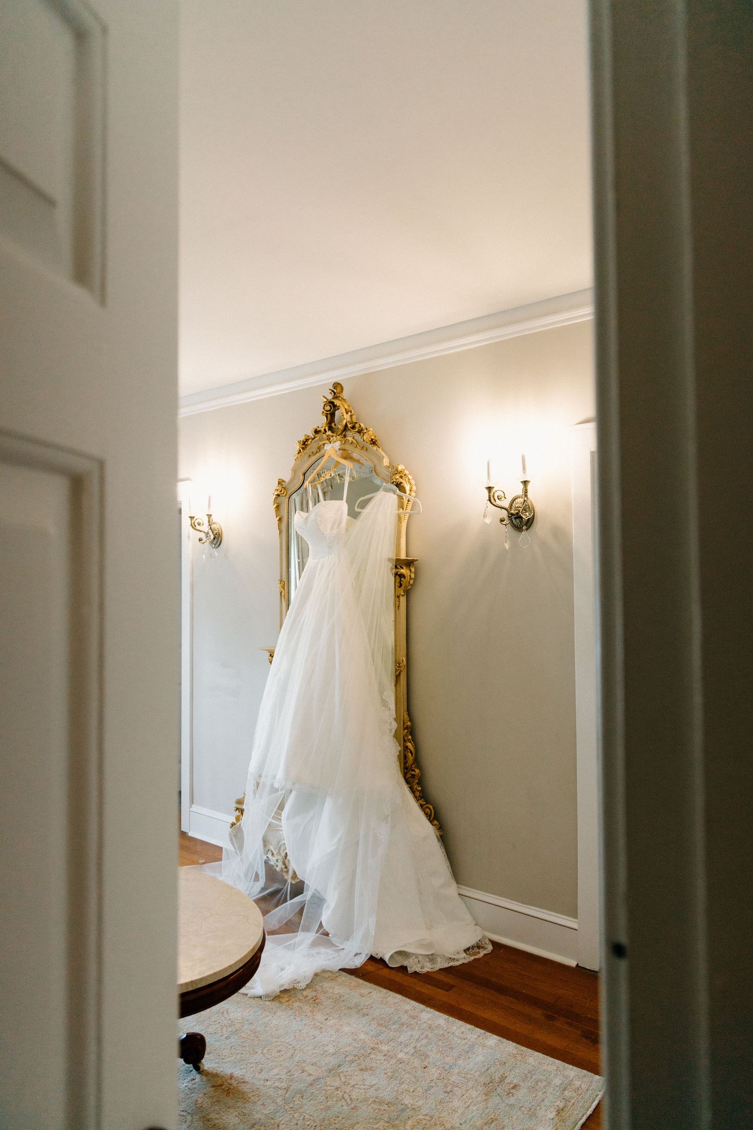 A white wedding dress hangs elegantly on display in an ornate room with gold-framed mirrors and wall sconces.