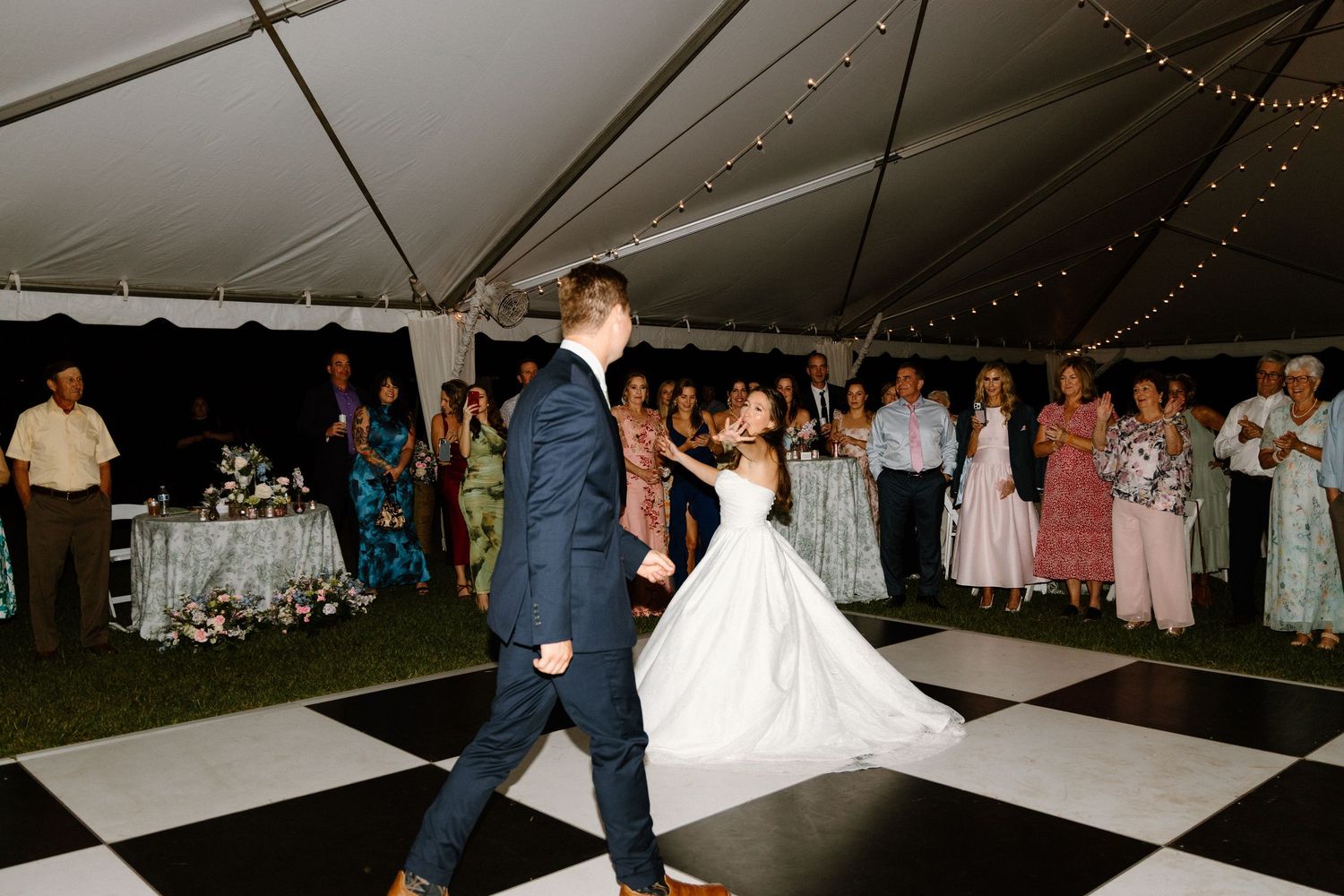 Couples dance together on a wooden floor under a white tent at an evening wedding reception.