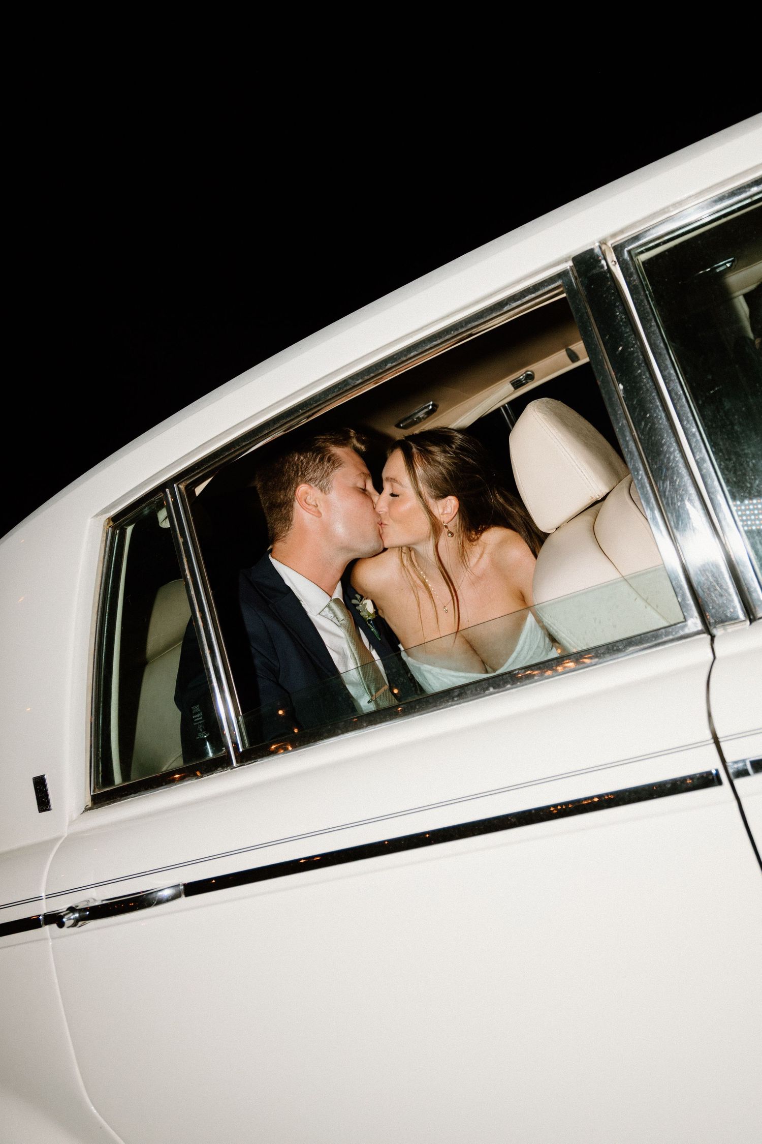 A romantic couple shares a kiss in the backseat of a white luxury car during what appears to be a wedding celebration.