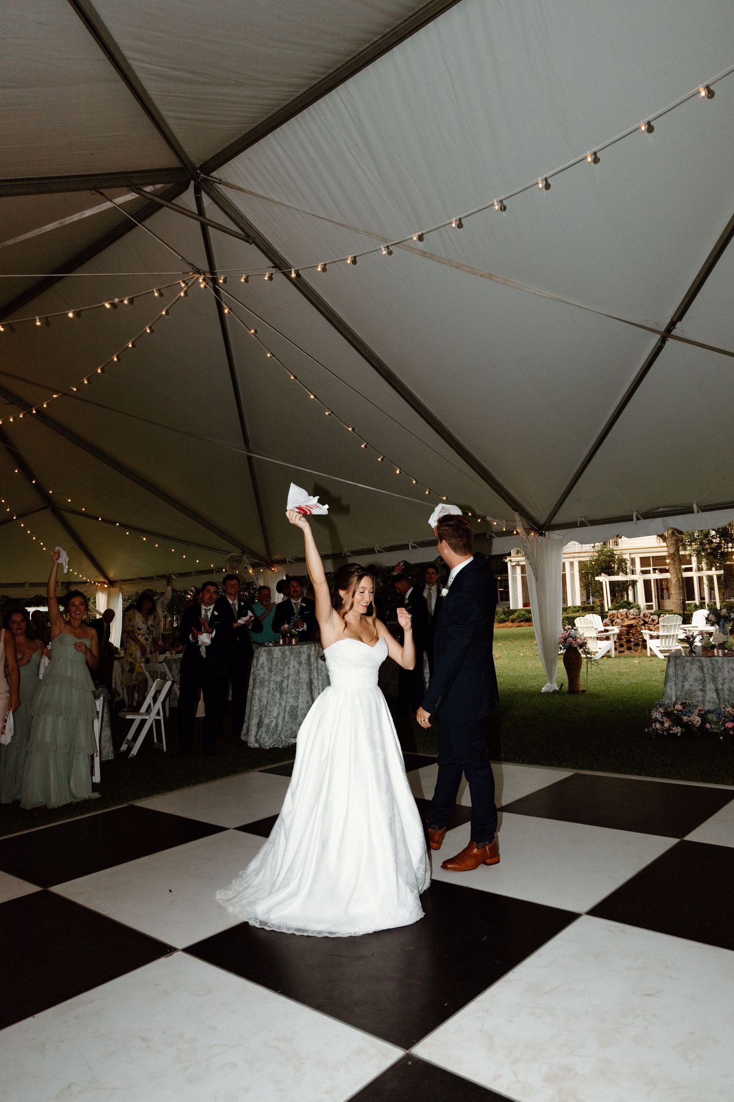 A couple dances together on a black and white checkered dance floor under a white tent with string lights overhead.