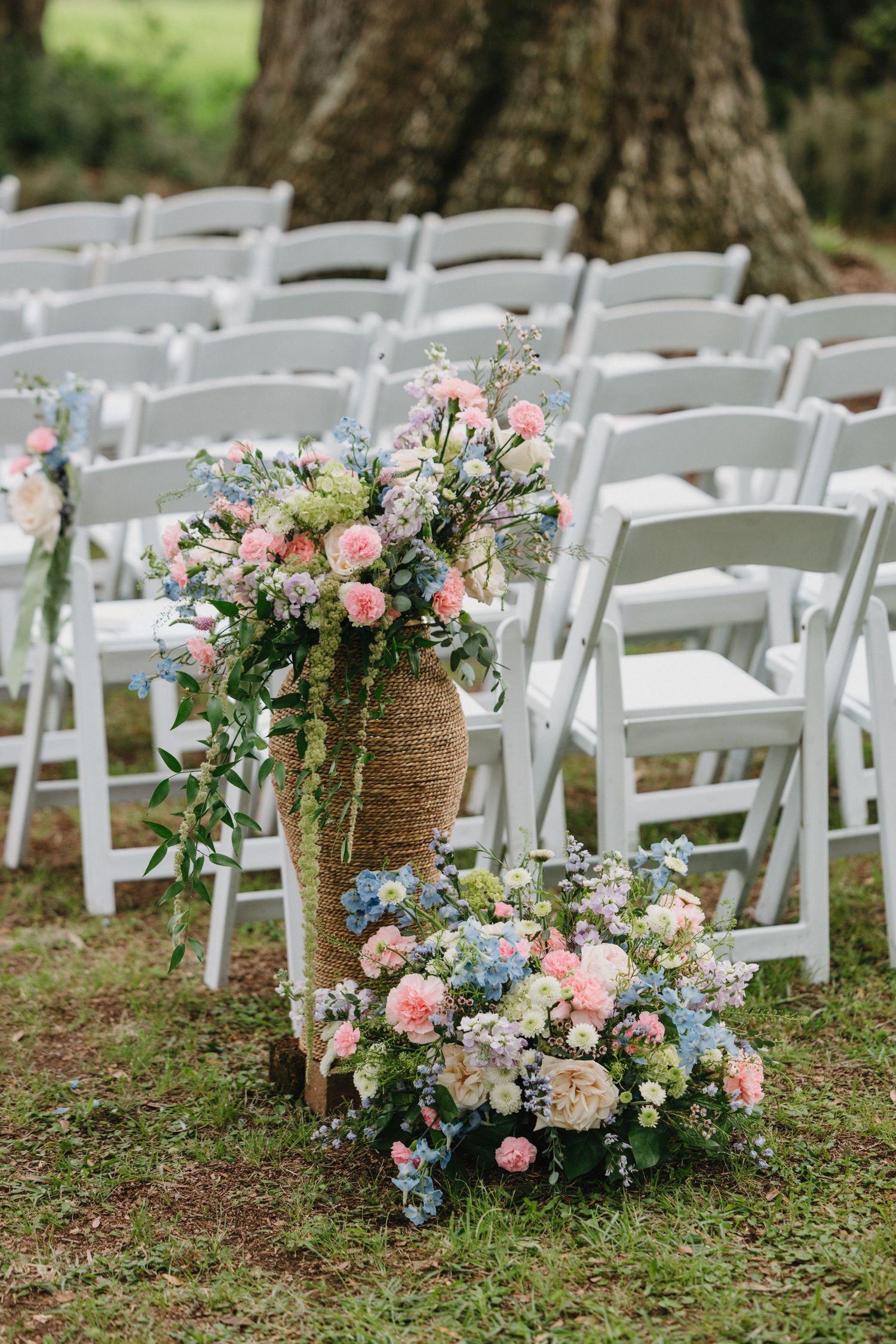 White folding chairs lined up for outdoor wedding ceremony with rustic burlap-wrapped floral arrangements along the aisle.