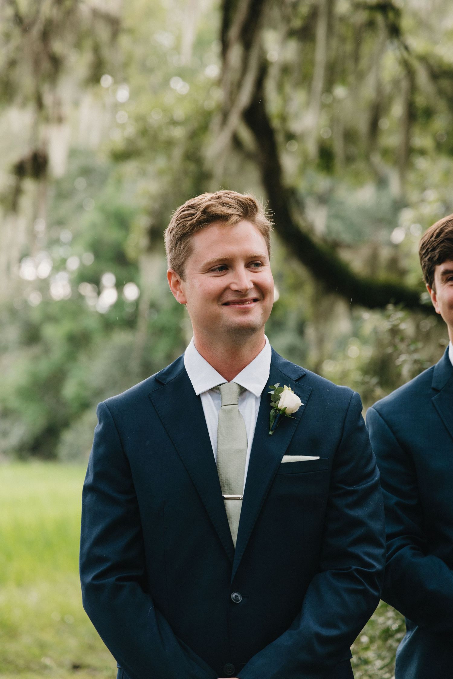 Groomsmen in navy suits with light colored ties smile during an outdoor wedding ceremony in a garden setting.