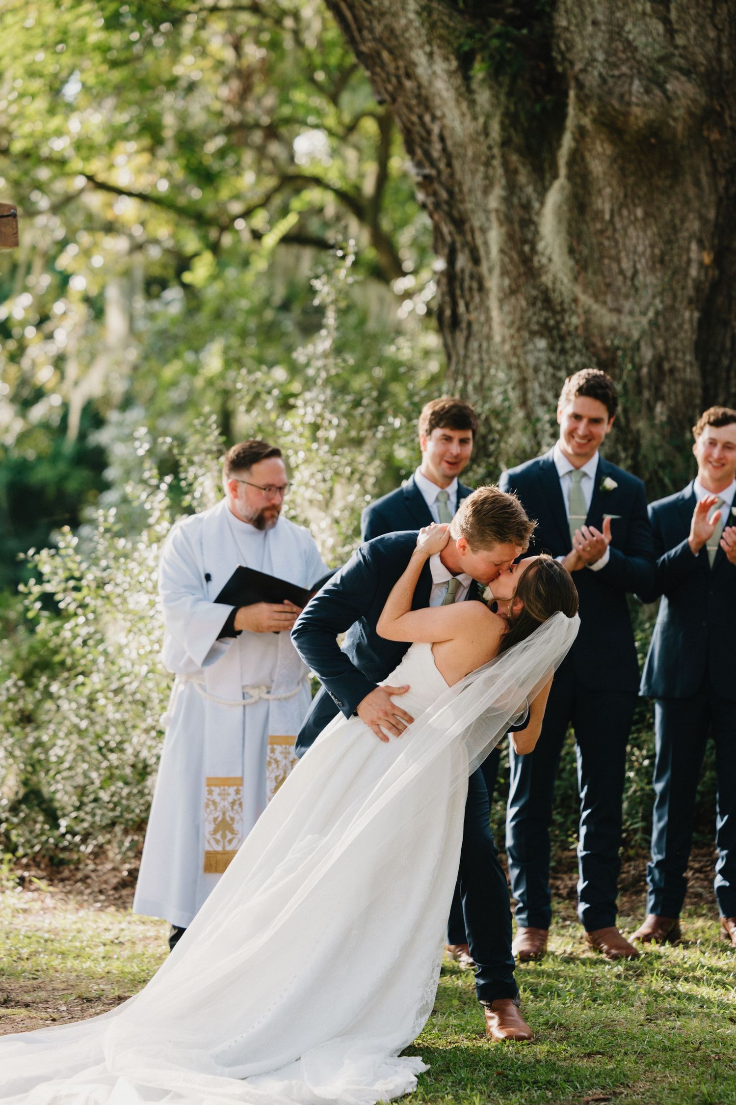 A romantic dip pose sequence during an outdoor wedding ceremony under large trees.