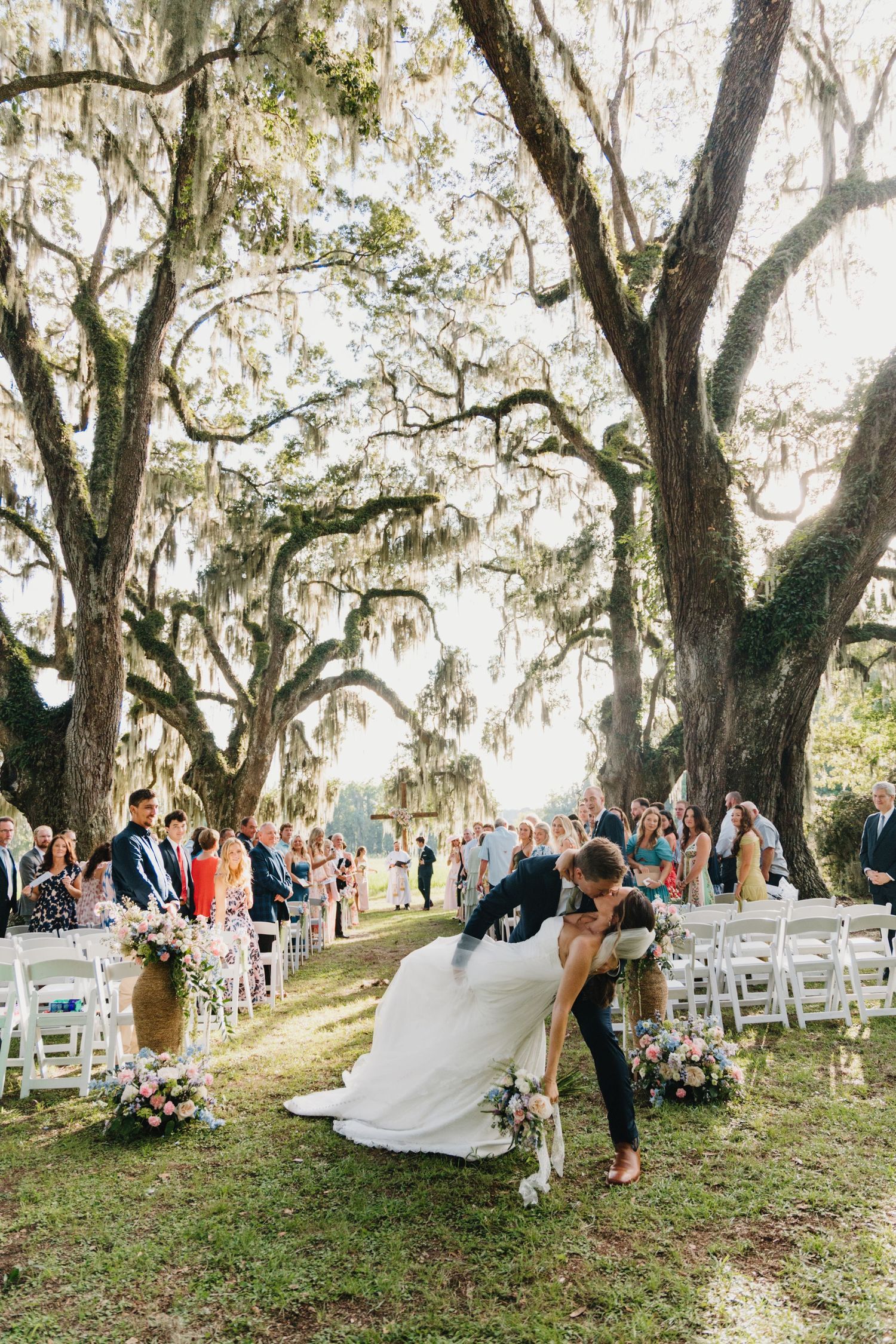 Wedding ceremony guests seated on white chairs under majestic oak trees draped with Spanish moss in an outdoor setting.