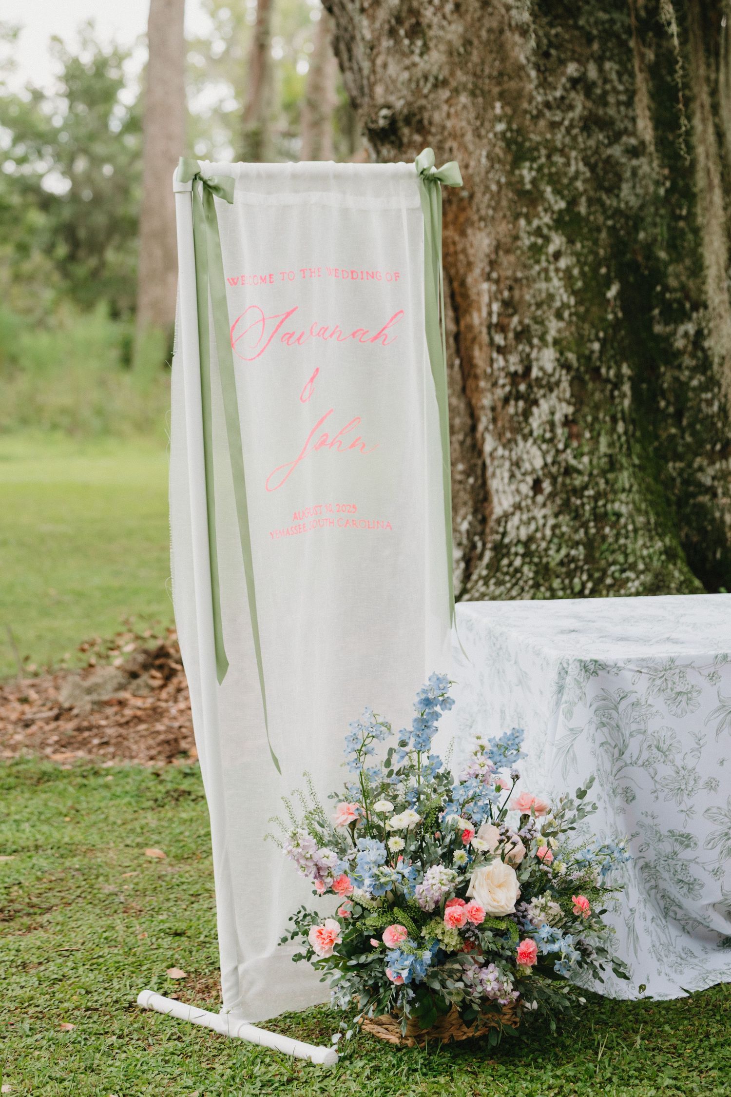 A white fabric banner with pink text hangs by a tree, accompanied by a delicate floral arrangement of blue and pink blooms.