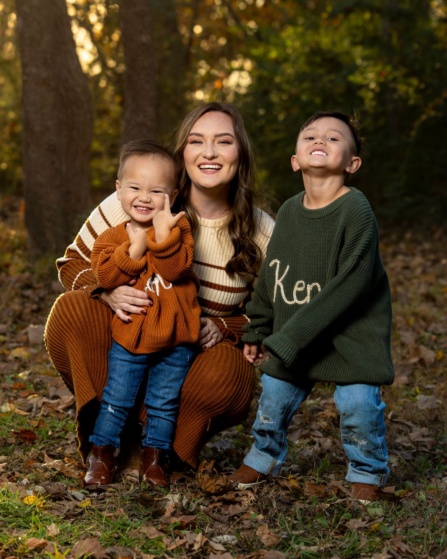 Three children sitting together in autumn leaves wearing cozy sweaters in brown and green tones in Fayetteville AR peak foliage.