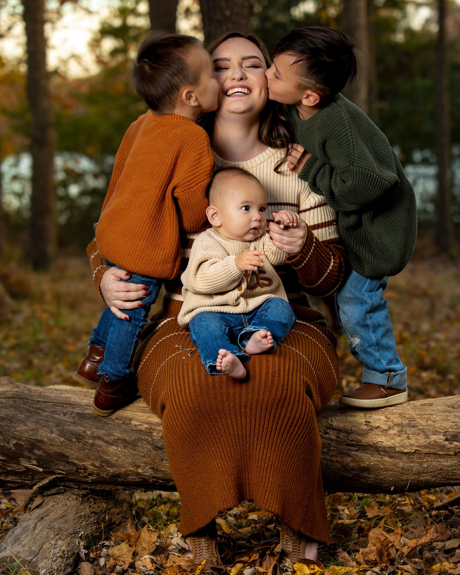 A family shares a joyful moment kissing mom on a fallen log in an autumn forest setting during golden hour in Fayetteville AR peak foliage.