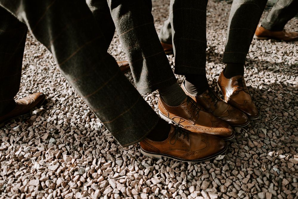 Brown leather dress shoes paired with dark pants on a textured gravel surface in natural lighting.