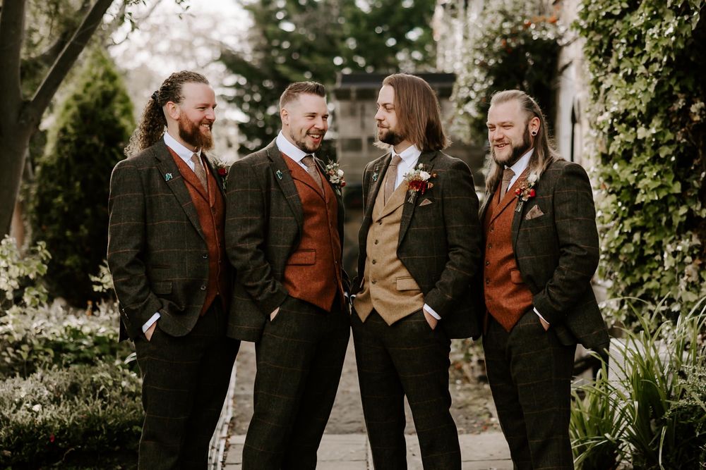Four groomsmen in dark suits with rust-colored vests share a candid moment in a garden setting.