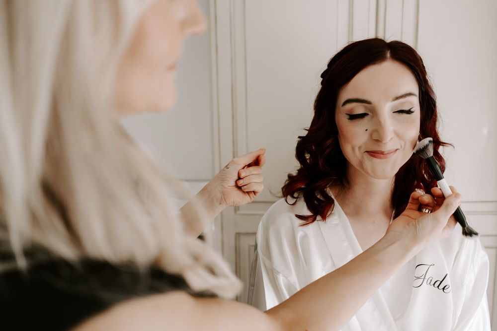 A bride in a white robe smiles while getting her hair and makeup done during wedding preparations.