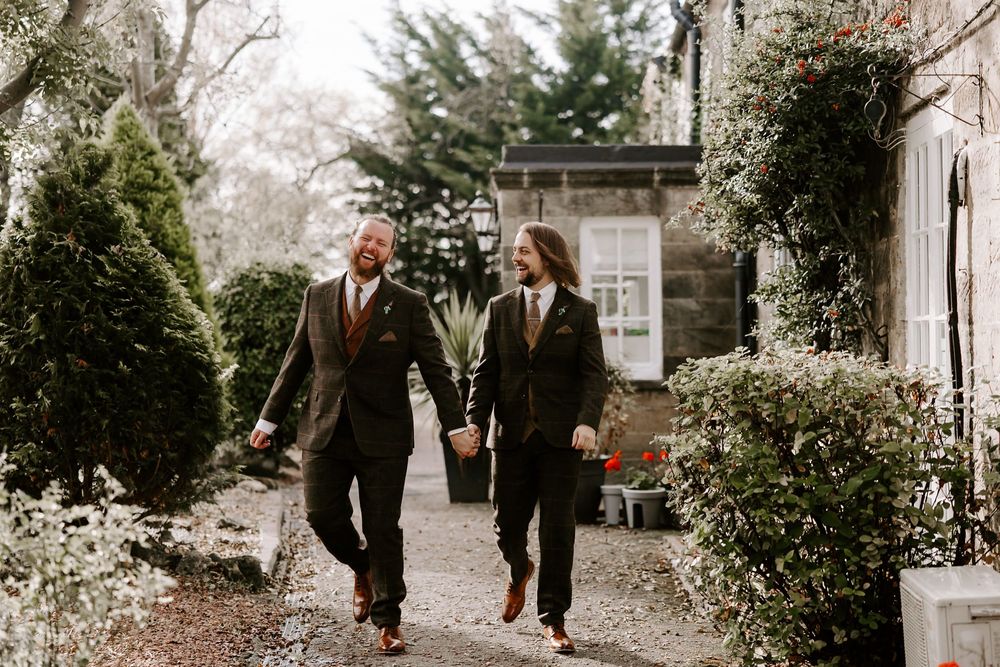 Two people walk hand in hand down a garden path between evergreen shrubs and vines on a cloudy day.