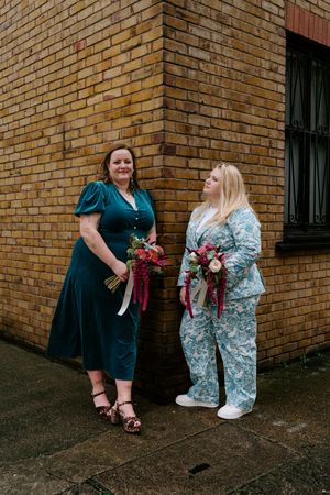 Two people pose with burgundy bouquets against a brick wall exterior.