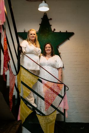 Two people in white dresses pose playfully with colorful fabric strips in an industrial-style room.