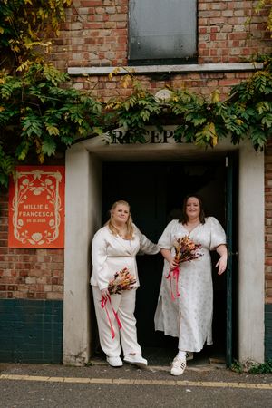 Two people in white outfits sit on steps near a brick wall with climbing vines and signage.
