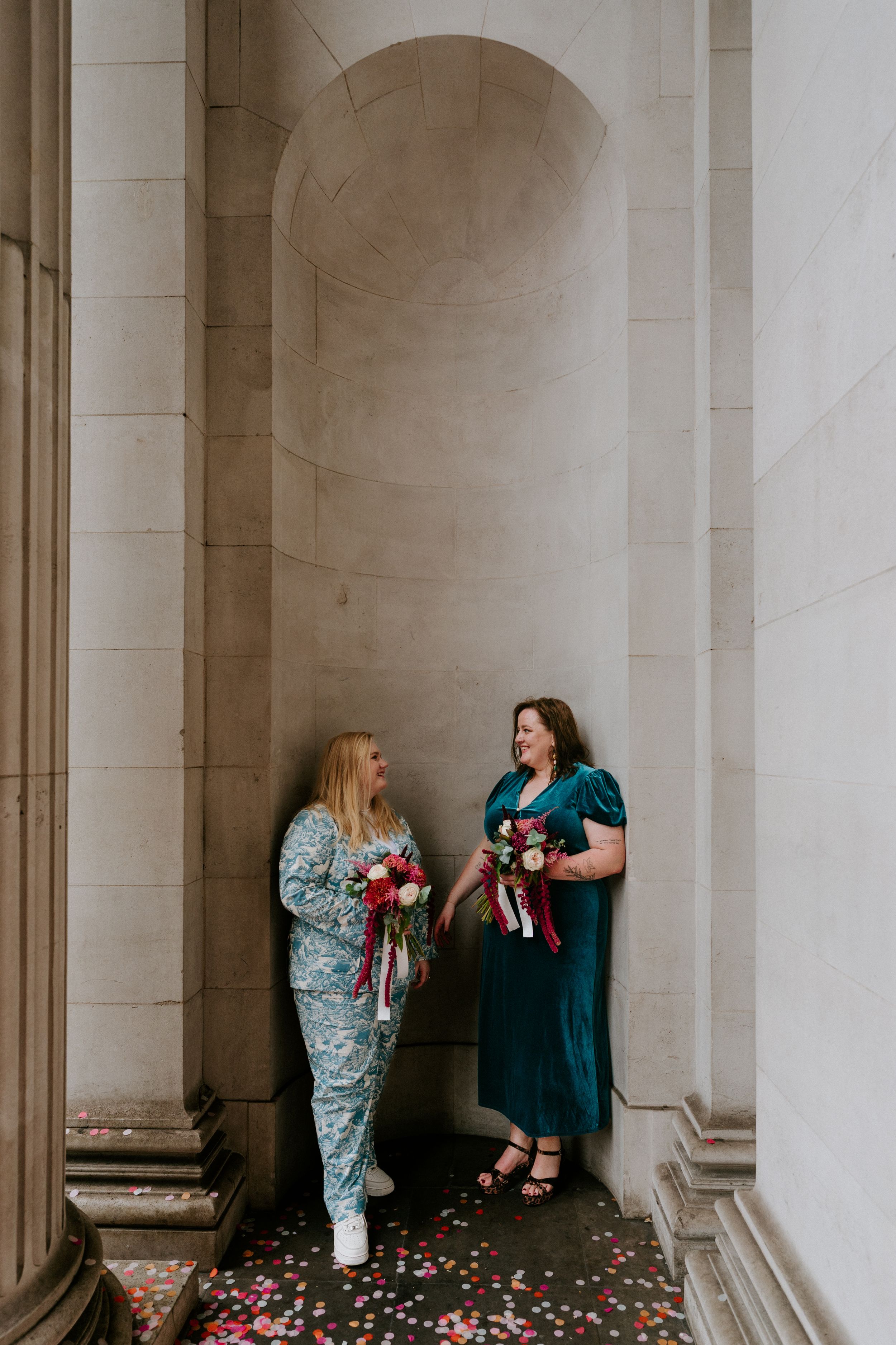 Two people stand in a classic columned hallway with white pillars while scattering rose petals on the ground.
