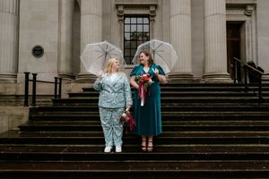 A wedding party with umbrellas poses on stone steps outside a grand building in rainy weather.