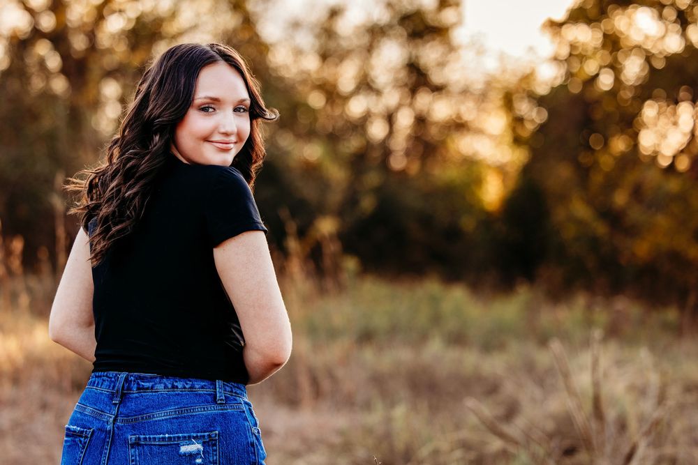 Woman in black top and blue jeans poses in a golden field during sunset.