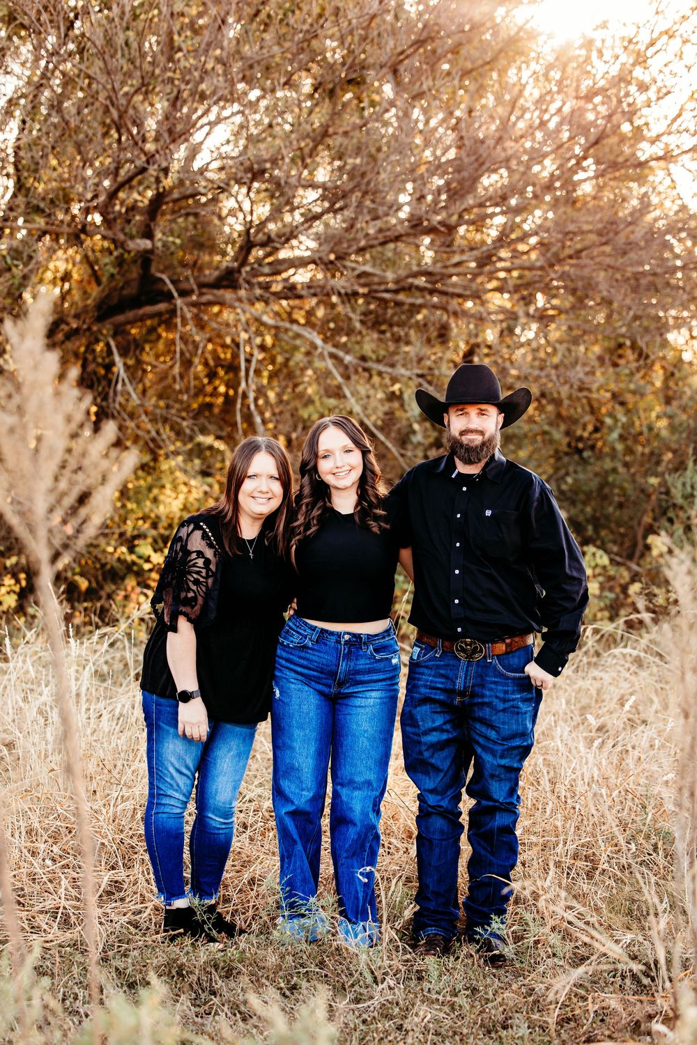 A family of three poses together in a rustic autumn setting with tall grass and trees in the background.