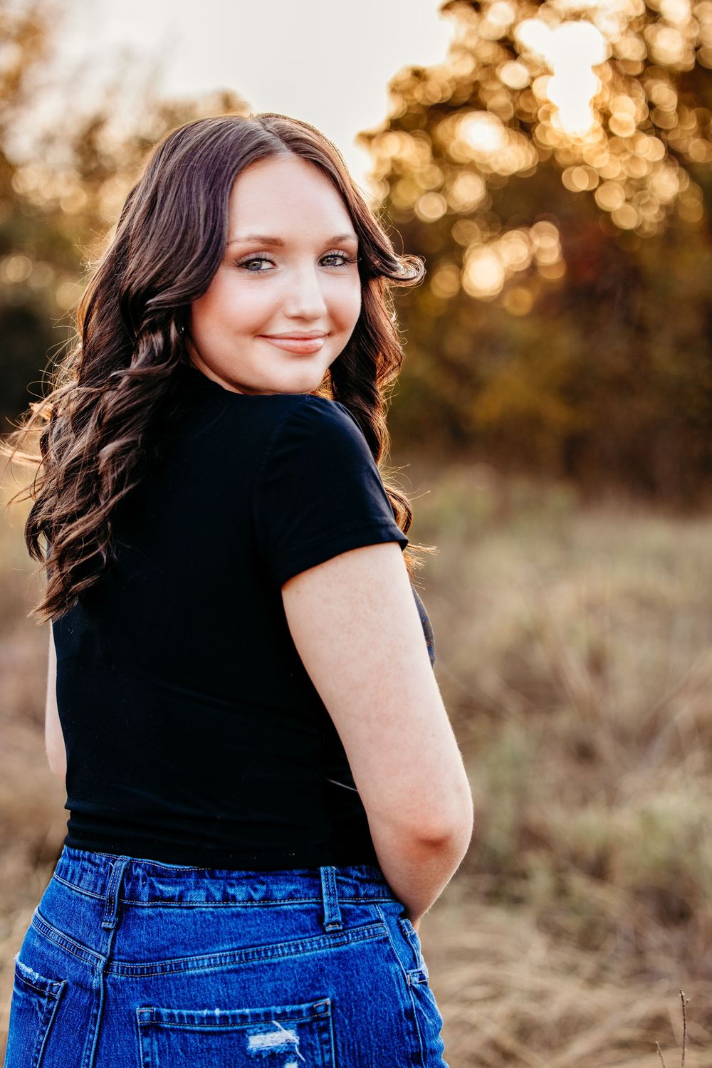 Woman in black top and blue jeans poses outdoors during golden hour sunset in nature setting.