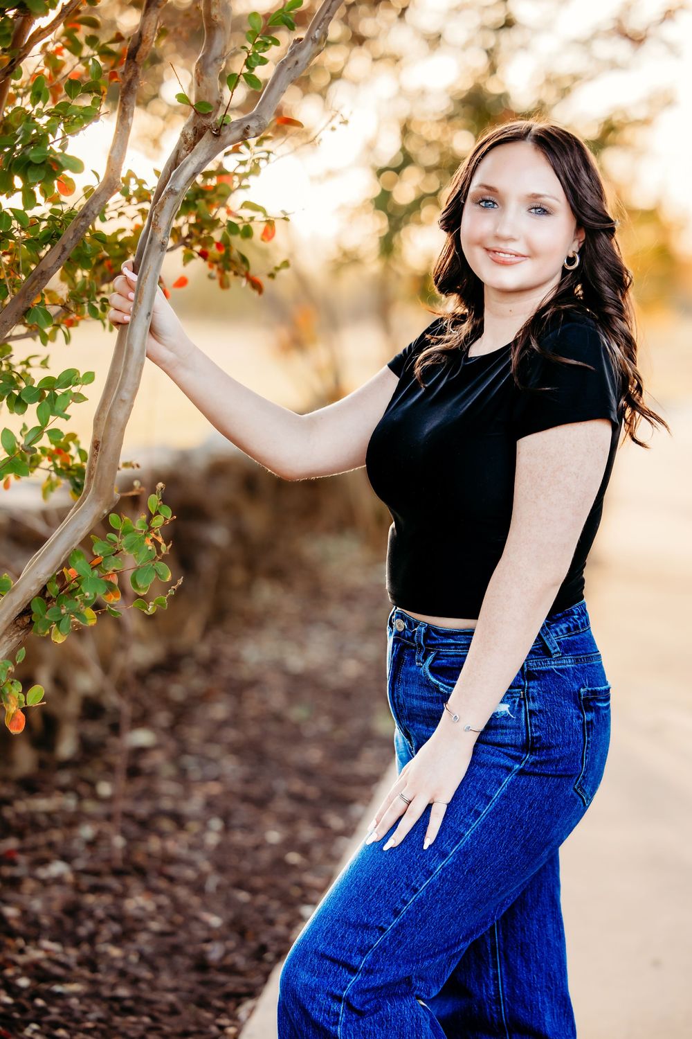 Portrait of someone in a black top and blue jeans posing outdoors near a tree with warm autumn lighting in the background.