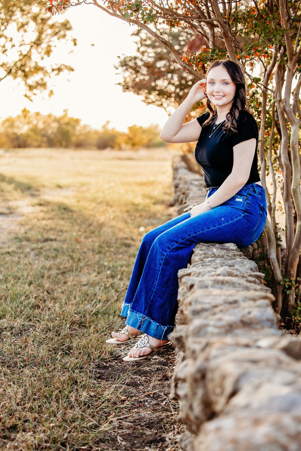 Person in blue jeans and black shirt sits on stone wall at sunset with autumn trees in background.