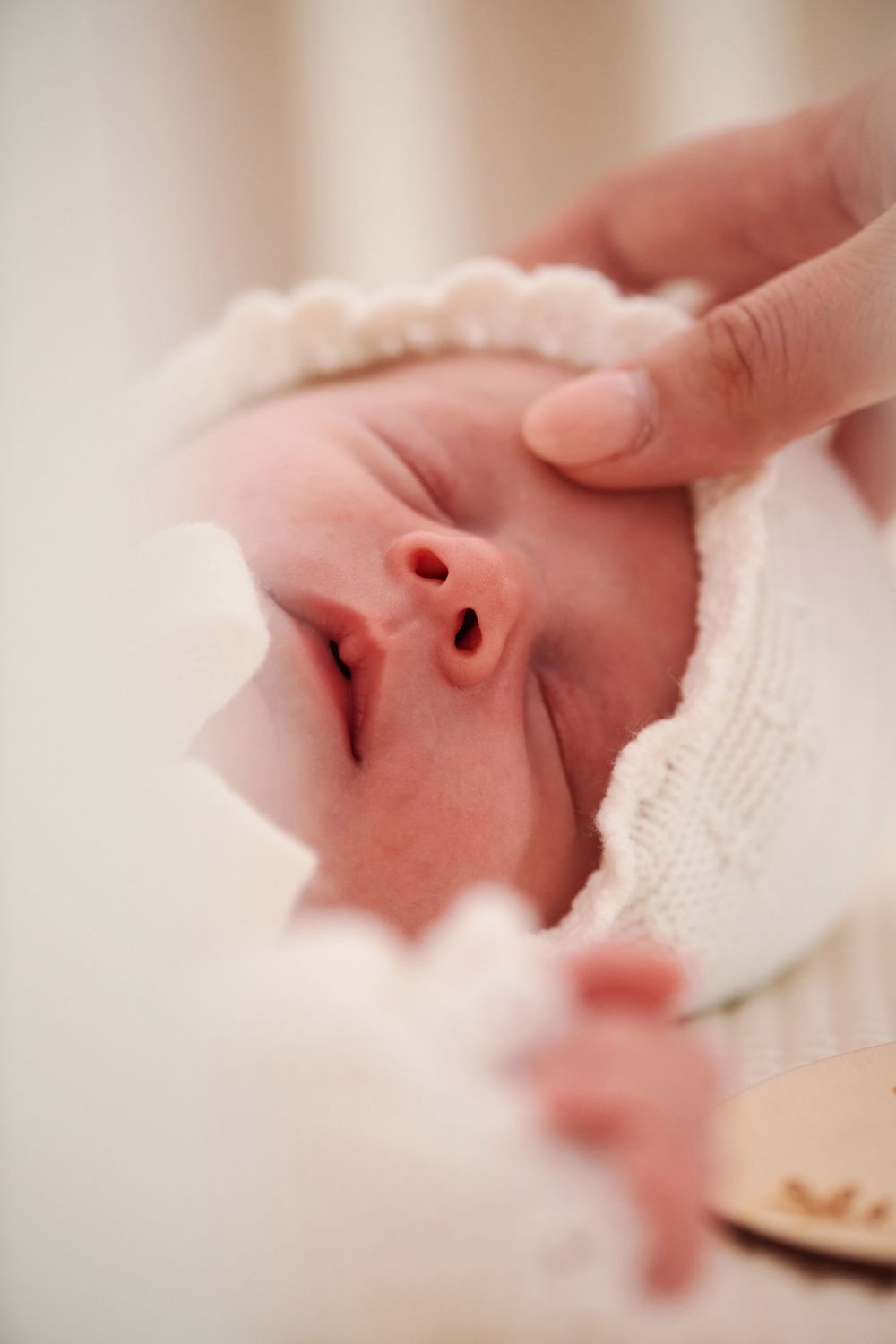 Close-up of a sleeping newborn baby wearing a white knit bonnet, gently touched by a parent.