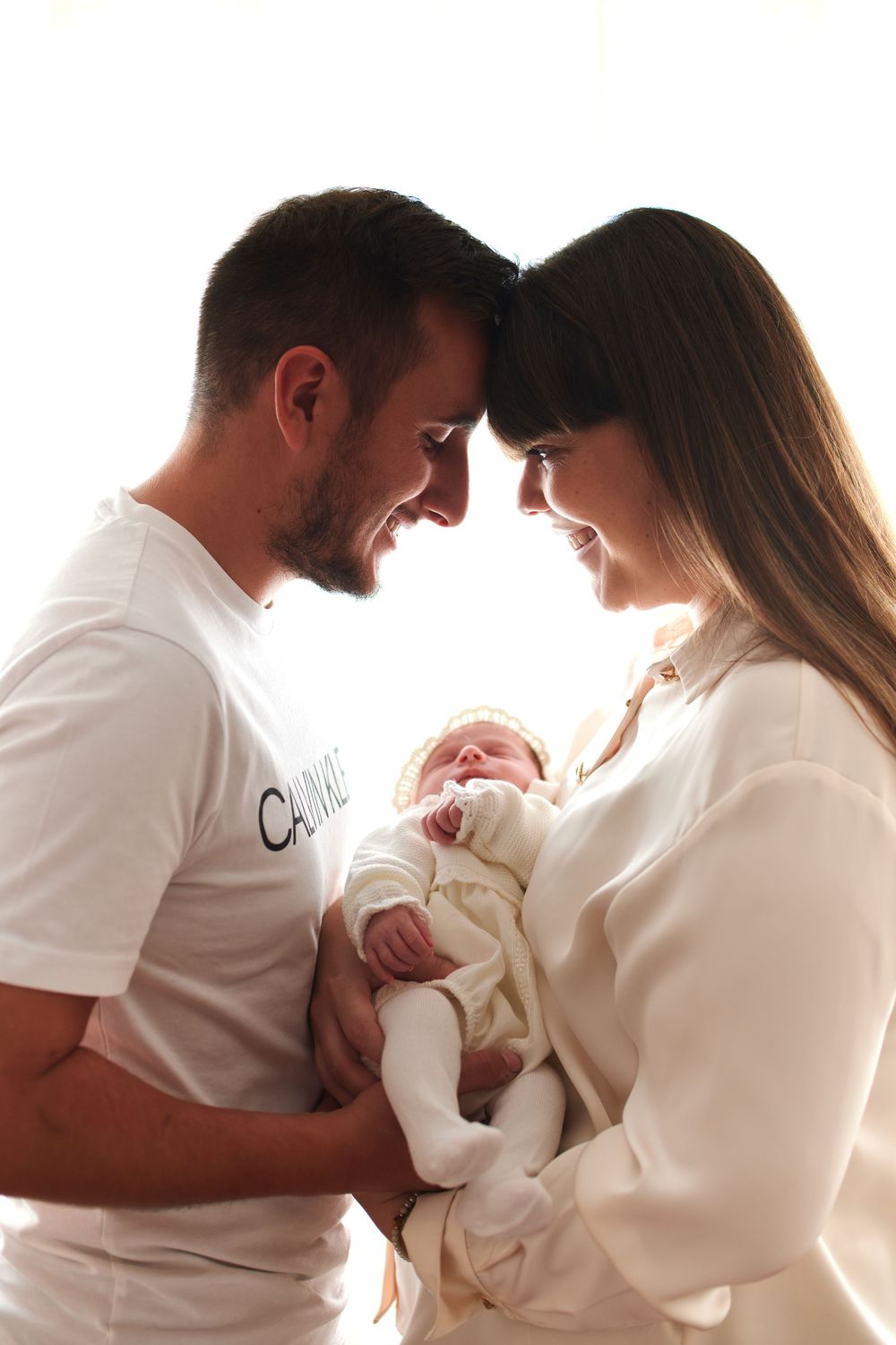 Loving parents holding their newborn baby together, smiling and touching foreheads in a bright room.