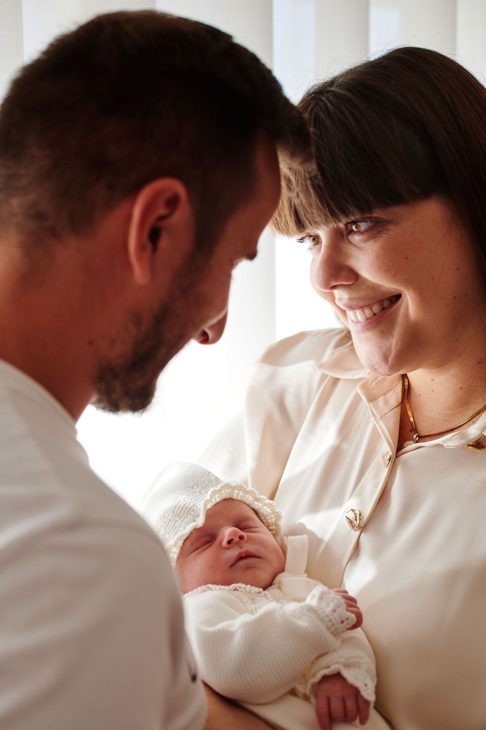 Smiling mother and father lovingly gazing at their newborn baby wrapped in a white blanket.