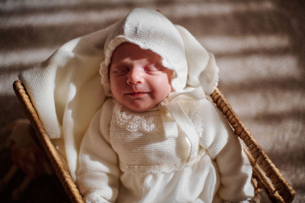 Newborn baby sleeping peacefully in a wicker basket, dressed in a white knit outfit and bonnet.