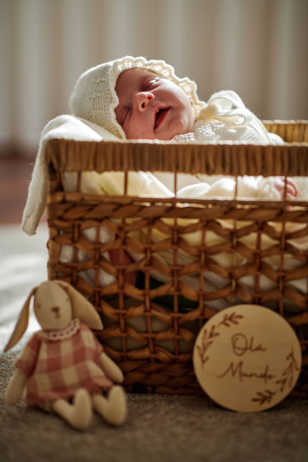 Newborn baby yawning in a wicker basket with a plush bunny and Ola Mundo wooden sign.