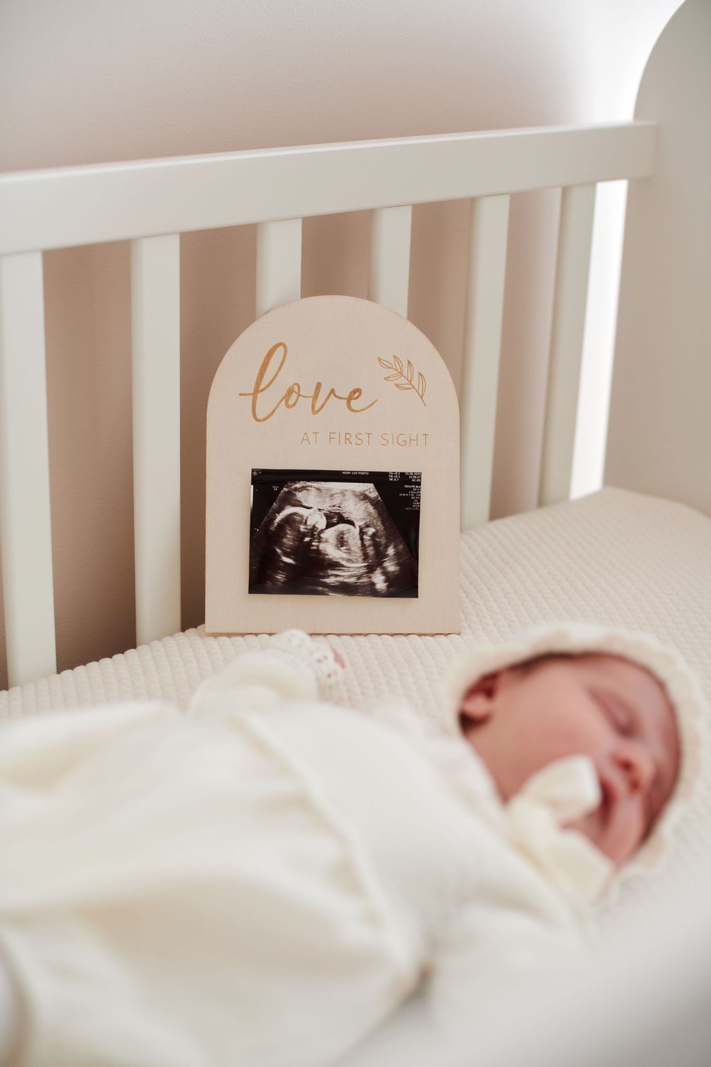 Newborn baby sleeping in a white crib beside a love at first sight ultrasound photo frame.