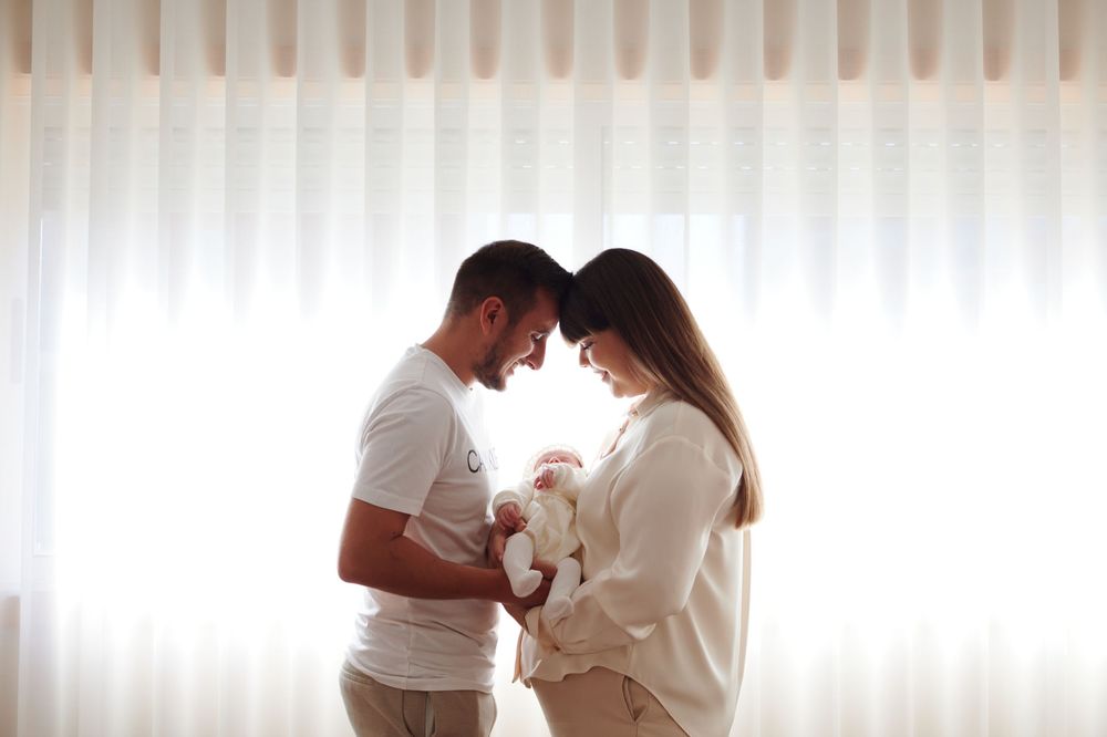 New parents standing by a bright window, tenderly holding their newborn baby together.