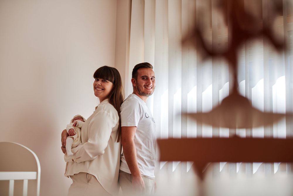 Smiling new parents standing back to back near a window in a bright nursery room.