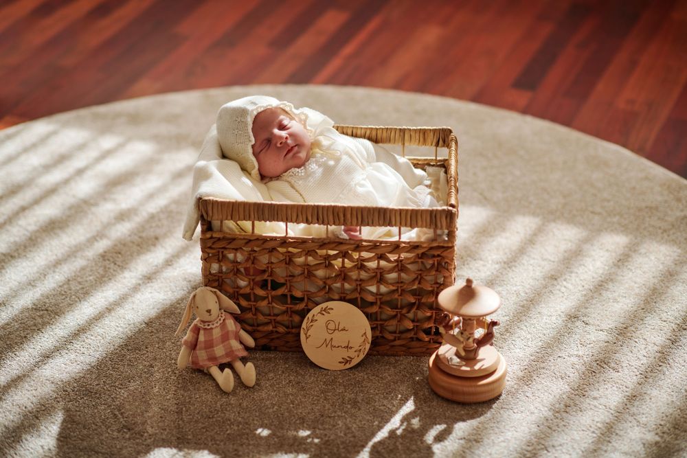 Newborn baby sleeping peacefully in a wicker basket on a rug with decorative toys.