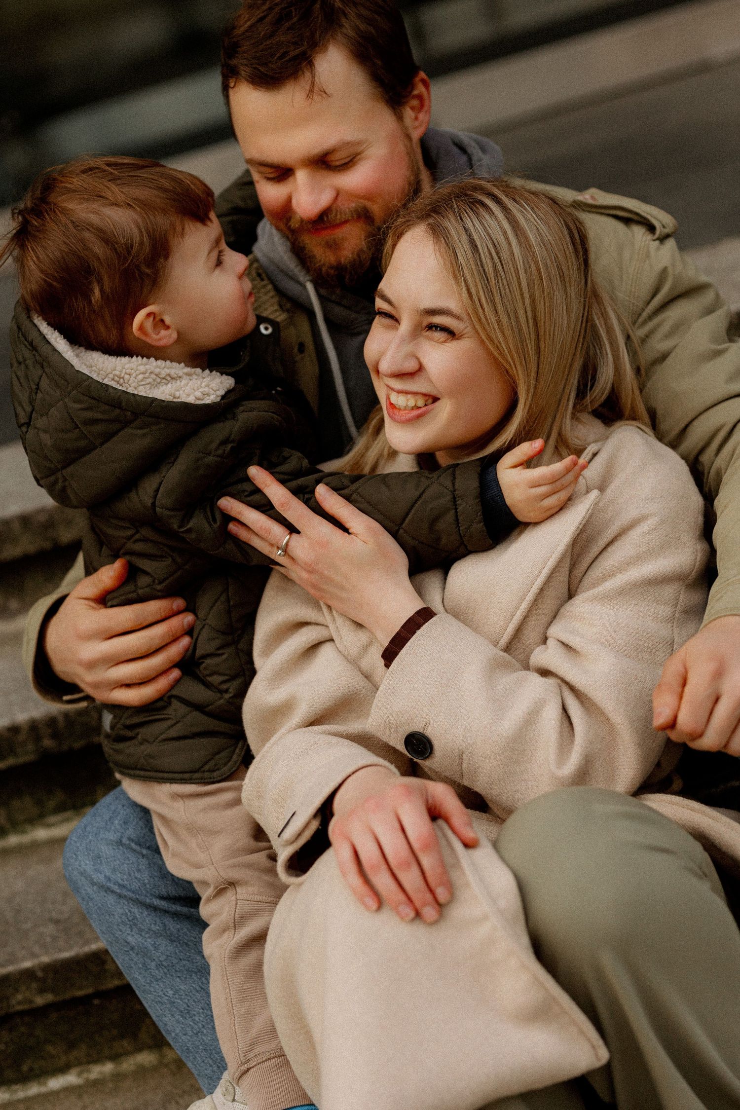 A couple shares tender moments with their young child while sitting together on steps in casual attire.