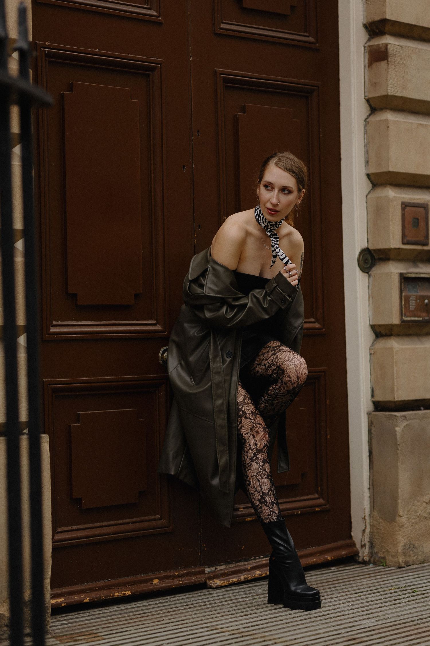 A person in black attire poses elegantly against a dark wooden door in a moody indoor setting.