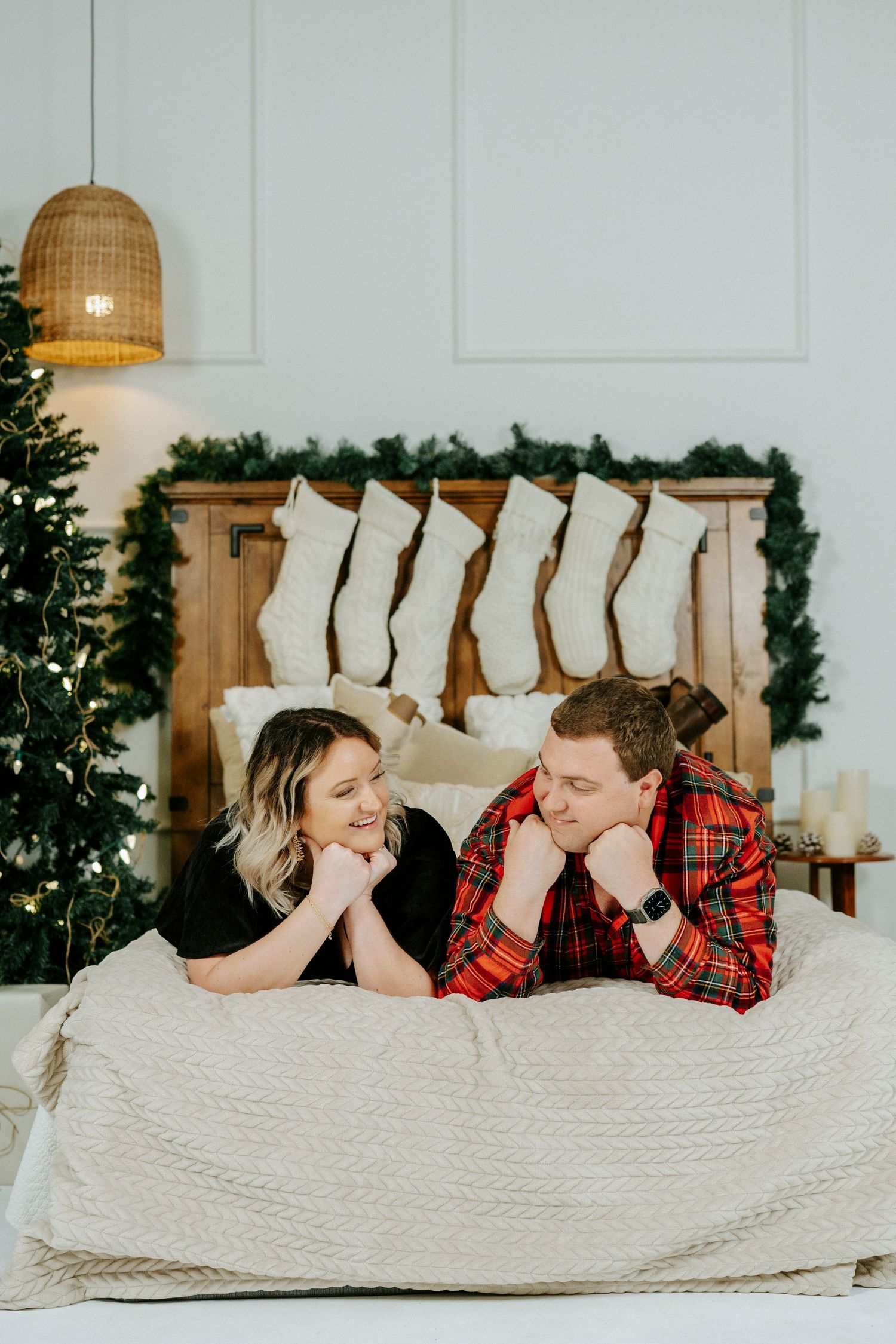 Couple lying on white bedding wearing matching red plaid shirts during cozy holiday photoshoot.