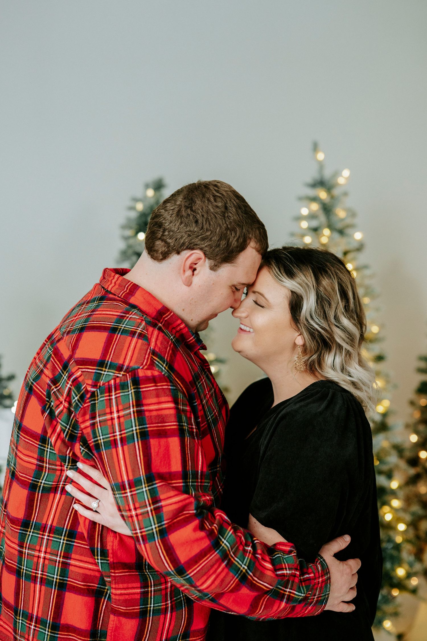 Couple in matching red plaid and black outfits embrace lovingly in front of lit Christmas trees.