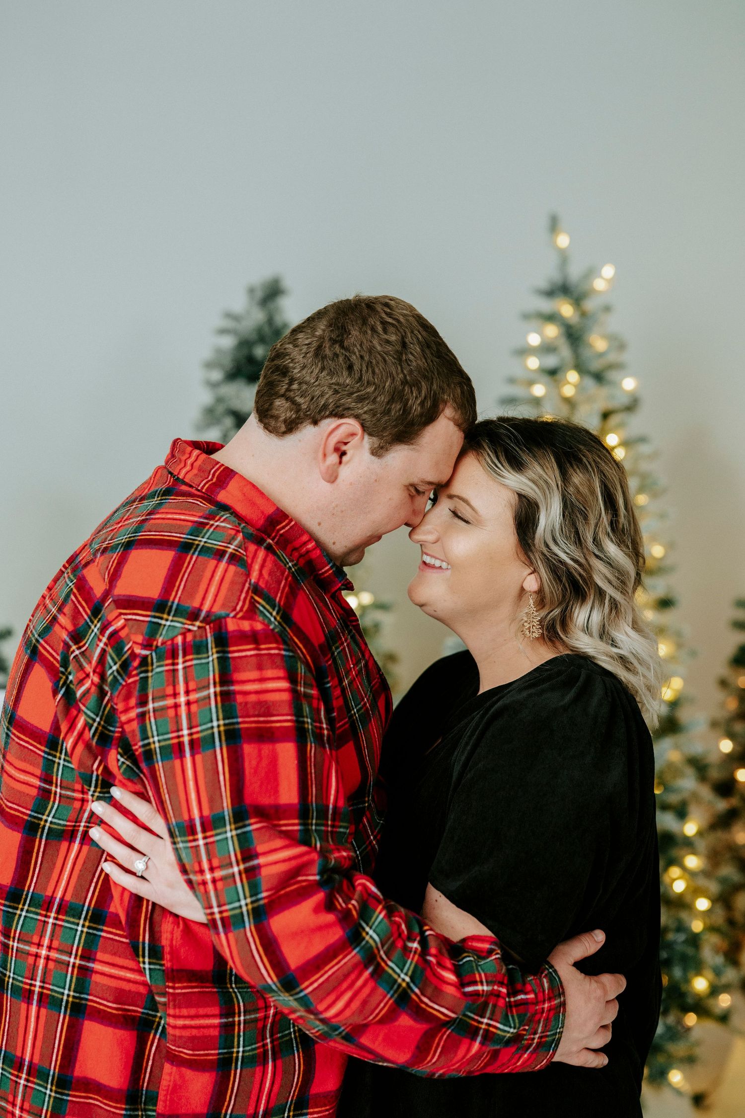 Couple embraces in front of Christmas tree with one wearing festive red plaid and the other in black.