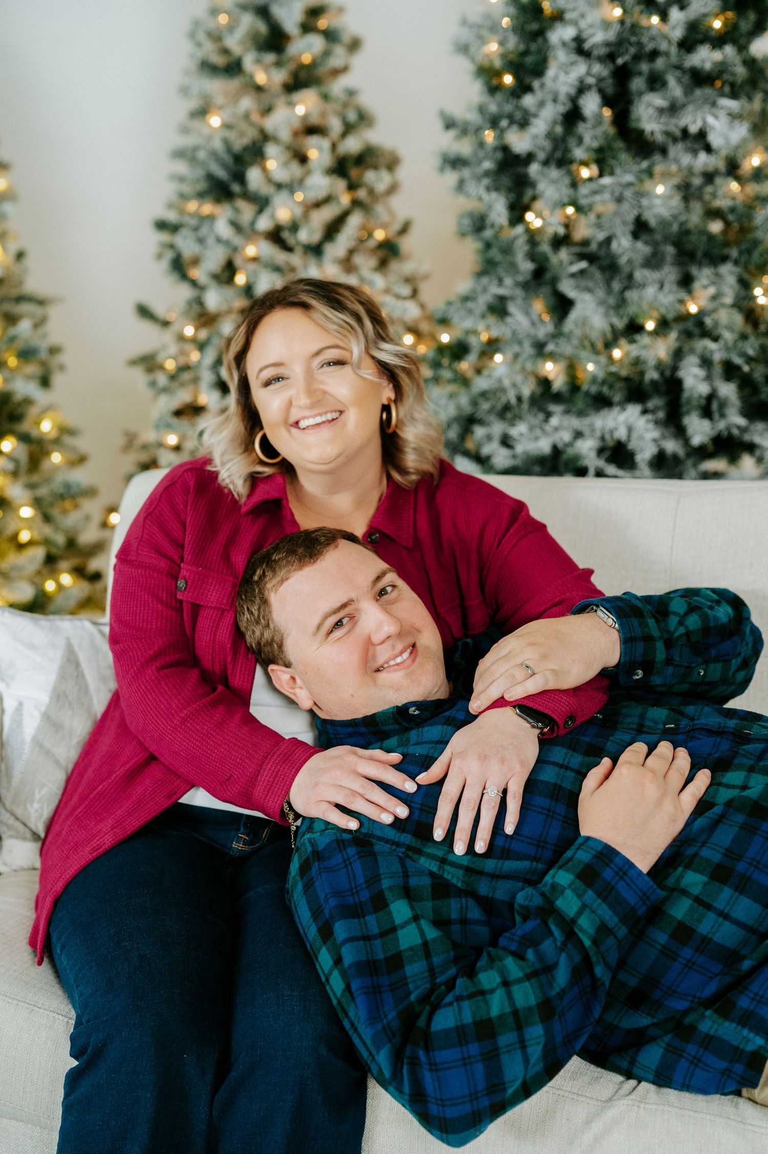 A couple in holiday attire laughs and embraces while posing for Christmas photos in front of a decorated tree.