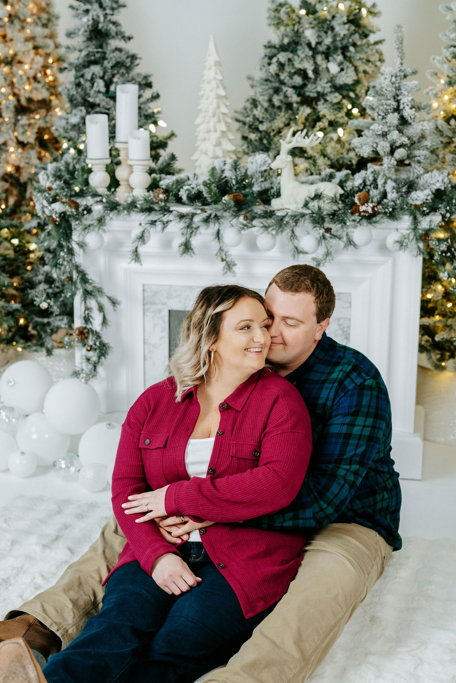 A couple in red and plaid sits together in front of a decorated Christmas mantel with white trees and holiday decor.