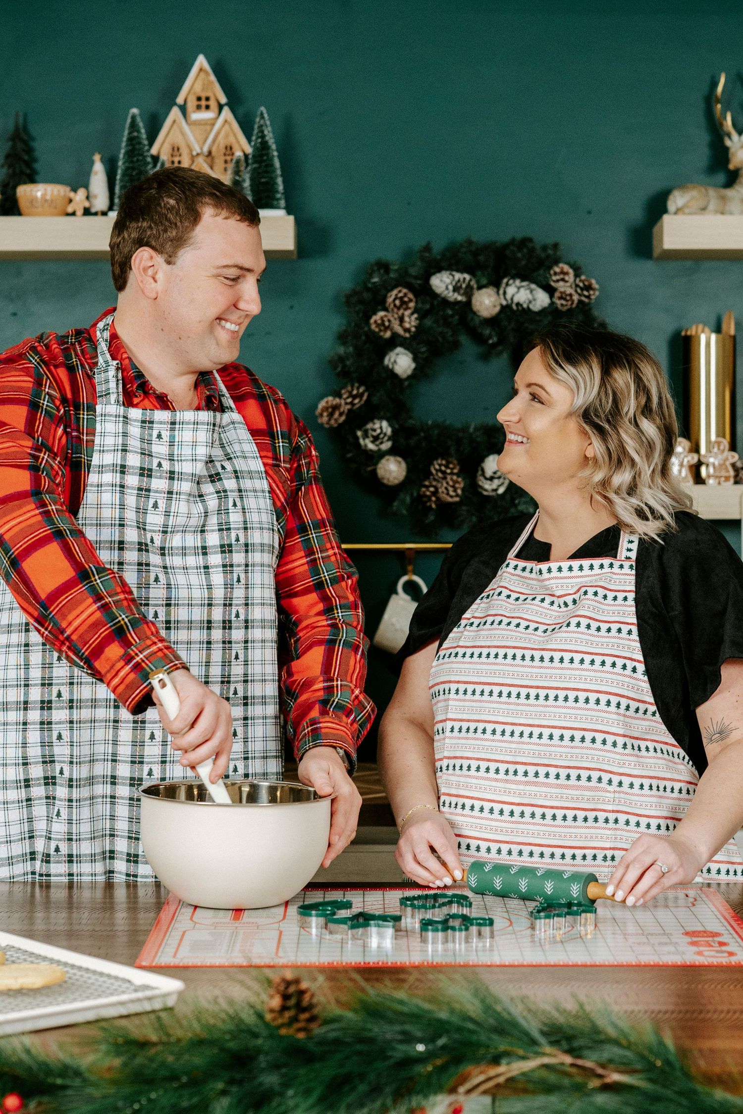 A couple in holiday attire shares a joyful moment while baking together in a festively decorated kitchen.