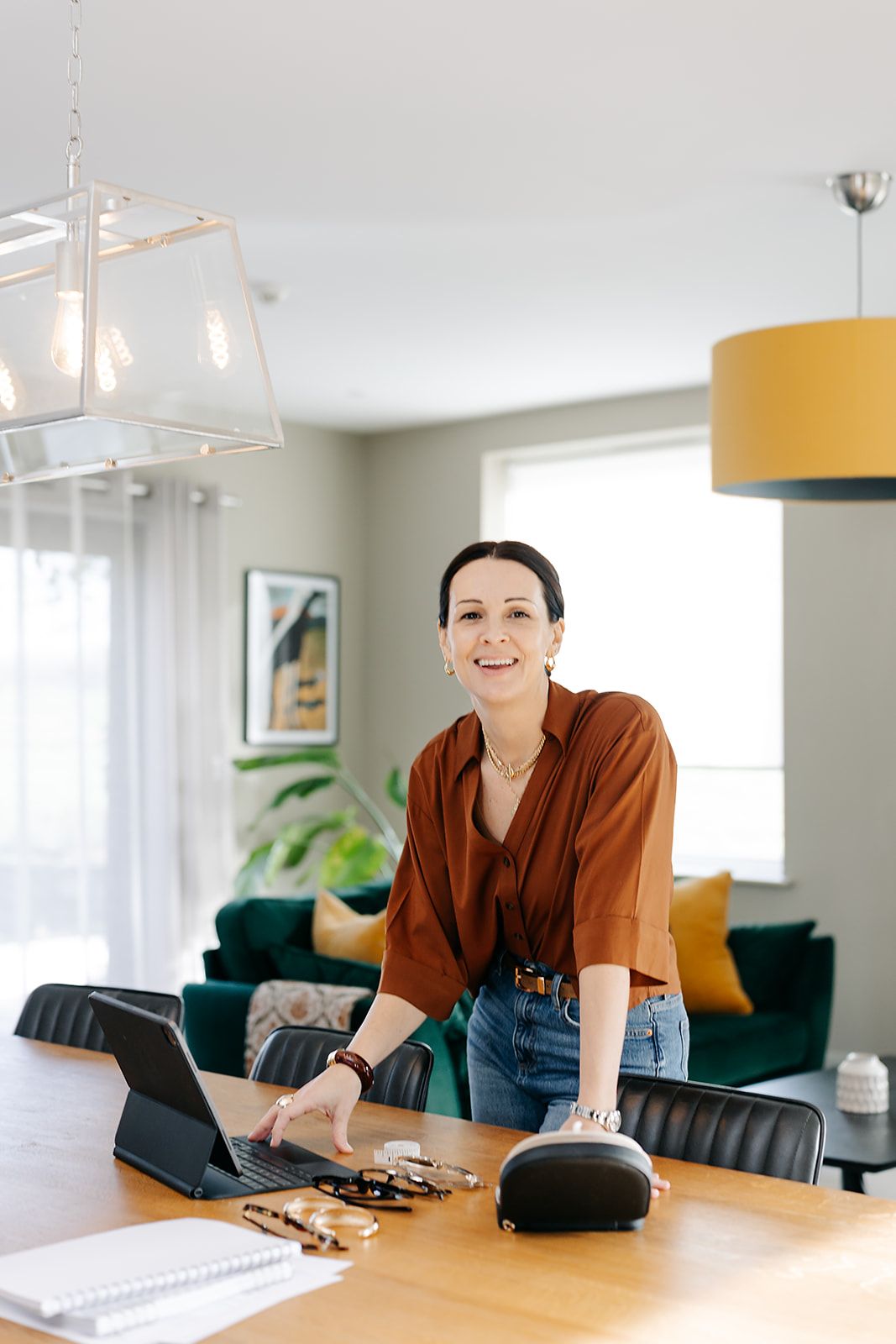 Person in brown shirt sits at modern office desk with yellow pendant lights and green plants in background.