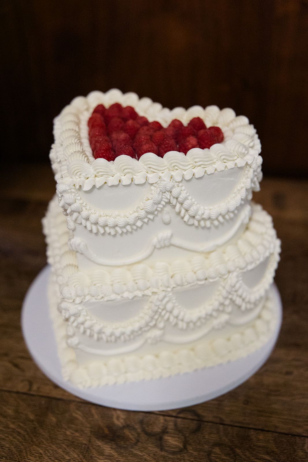 A white wedding cake decorated with delicate pearl piping and topped with fresh red raspberries on a rustic wooden surface.